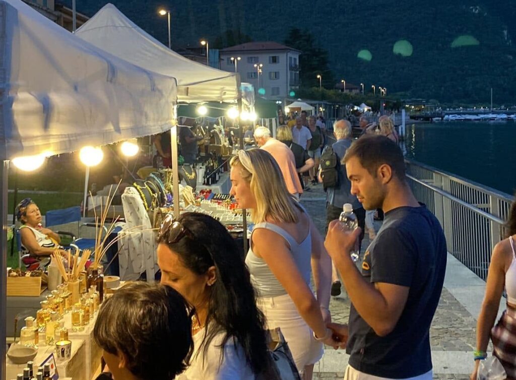Bancarelle illuminate del mercatino serale di artigianato sul lungolago di Porlezza al tramonto, sul Lago Ceresio
