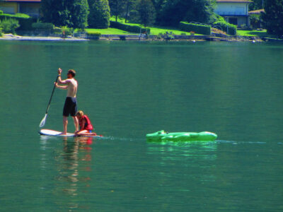 Stand-up paddle sul Lago Ceresio: adulto e bambino pagaiano sulle acque calme davanti alla riva verde di Porlezza